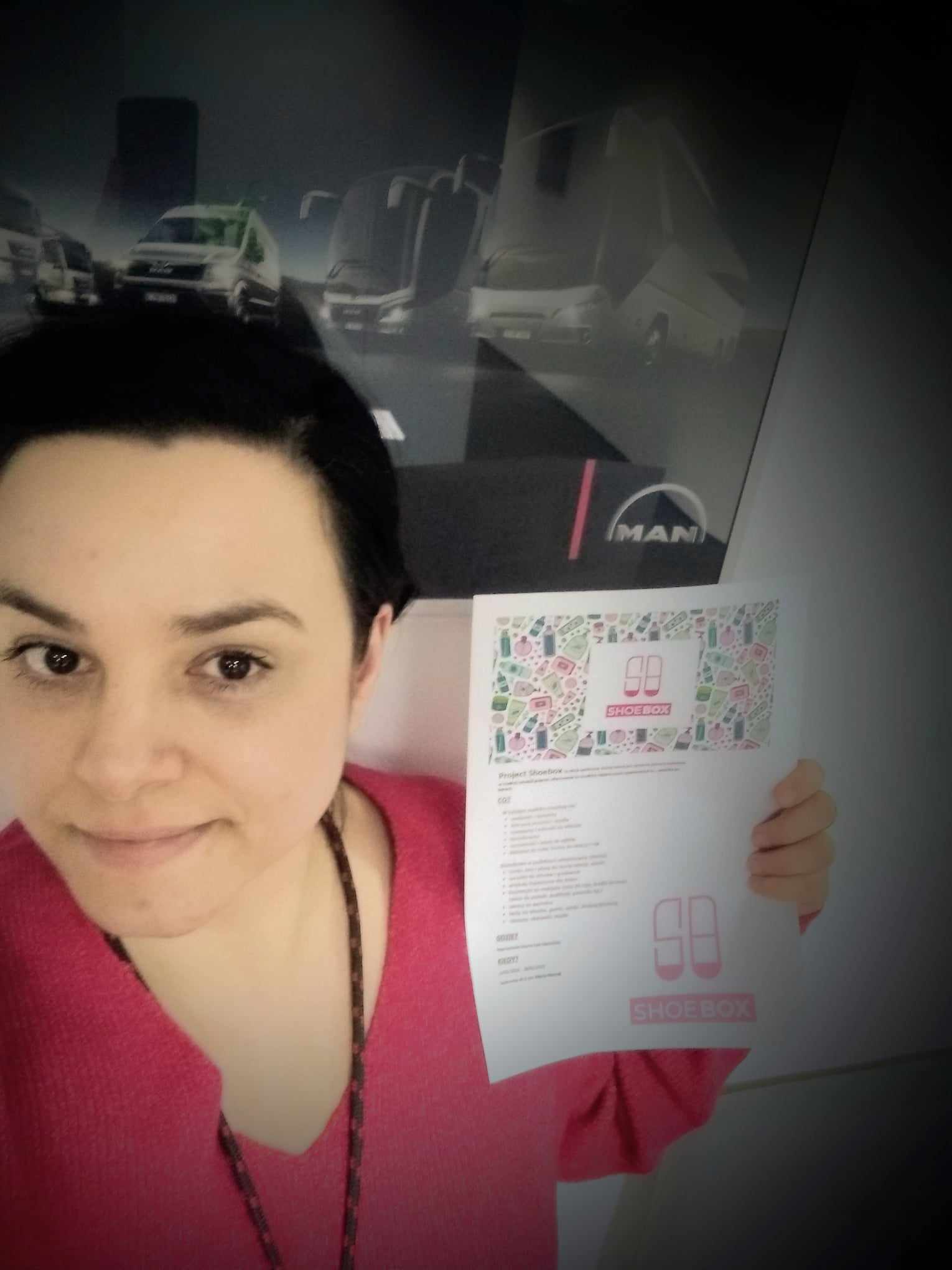 Młoda kobieta trzyma w ręku kartkę z napisem shoe box. Znajduje się w pomieszczeniu biurowym.